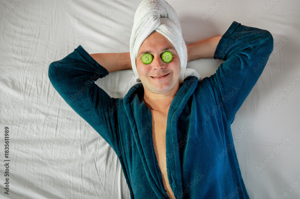 Fototapeta premium Man with cucumber eye mask in bathrobe, having fun during skincare