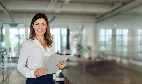 Happy 45 years old Latin professional middle aged business woman corporate leader, happy mature female executive, lady manager standing in office holding clipboard looking at camera, portrait. Banner