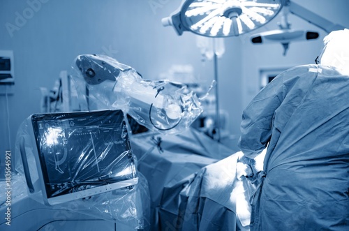 In a sterile Operating Room, a surgeon in scrubs uses a robotic system. The robotic arms hover over the patient, while the monitor displays vital information during surgery