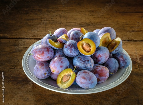 Damson plums, some halved to reveal golden flesh, are piled high on a decorative plate. Placed on aged wooden boards, the fruit evokes a sense of rustic charm