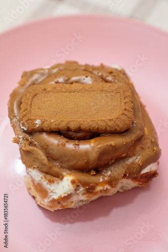 Vertical close-up of a freshly baked cinnamon roll smothered in creamy biscoff spread and topped with a whole lotus biscoff cookie on a pink plate
