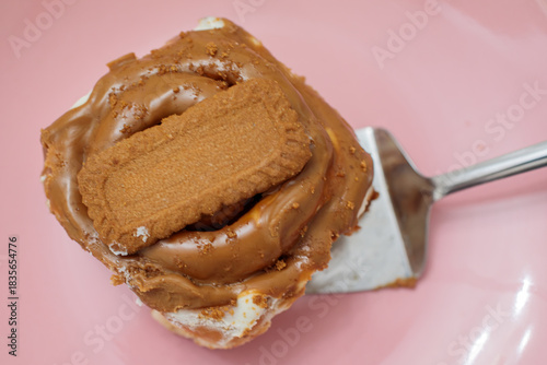 Freshly baked cinnamon roll topped with creamy biscoff spread and a lotus cookie, being served on a metal spatula on a glossy pink plate