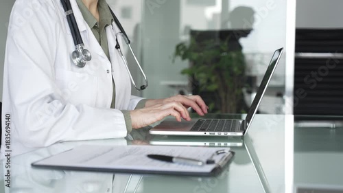 Healthcare female professional in white coat with stethoscope typing on laptop, managing patient records and telehealth data at desk. Medicine, health care and science concept