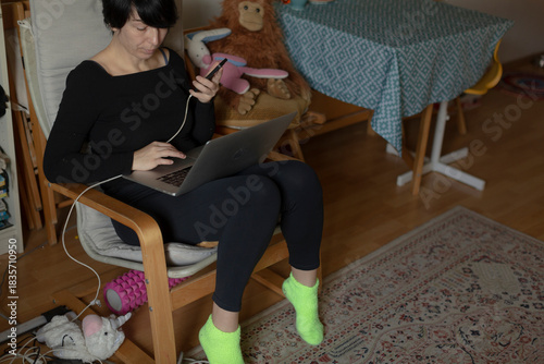 woman on chair with phone, tablet. multitasking parent in casual outfit scrolls apps while wearing neon socks, surrounded by teddy, drying rack and scattered toys, livingroom carpet and bright colors,
