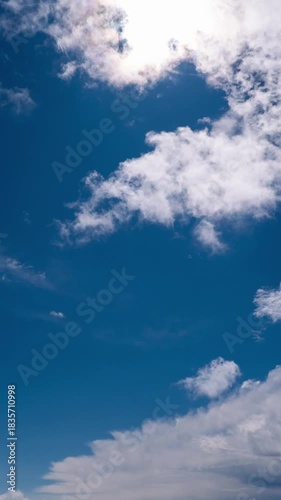 Vertical time lapse shows white puffy cumulus clouds drifting quickly across a vibrant deep blue sky. The bright sun shines prominently in a clear daytime atmosphere.