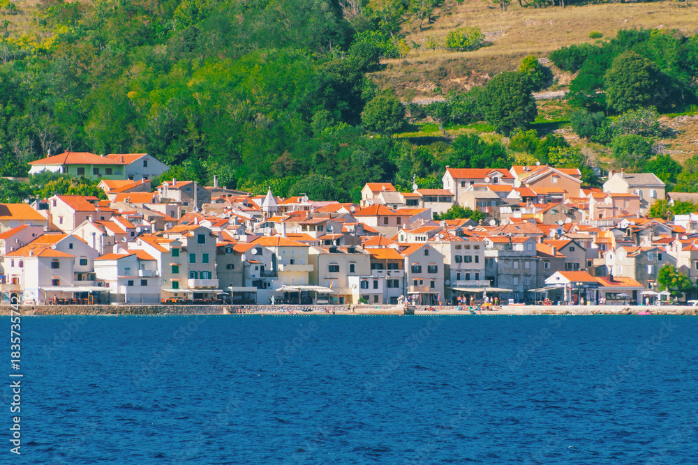 Obraz premium Picturesque coastal village with orange roofs and lush green hills by the sea. Baška, Krk island, Croatia