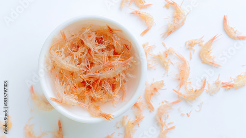 Dried small shrimp in white bowl with scattered shrimp shells on white surface, creating light and delicate seafood texture
