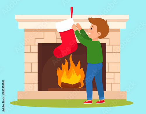 A young boy hangs a red and white Christmas stocking on a mantelpiece above a roaring fire.