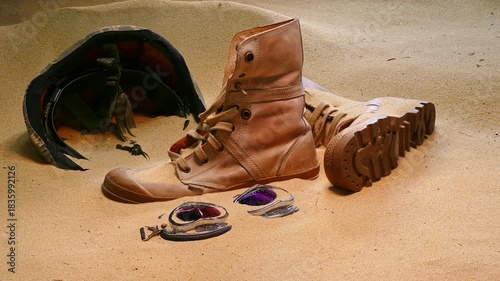 An arrangement of abandoned military gear, including a worn out boot, helmet, and shattered protective eyewear, partially buried in desert sand, symbolizing the harsh realities of conflict.