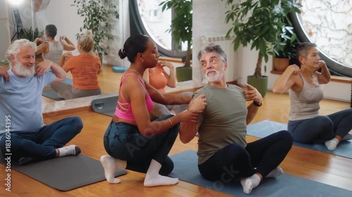 Black female fitness instructor guiding an older man through proper arm stretching during a senior group class. A group of seniors at a joint fitness class with a female trainer.