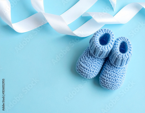 Blue knitted baby booties and white ribbon on pastel background