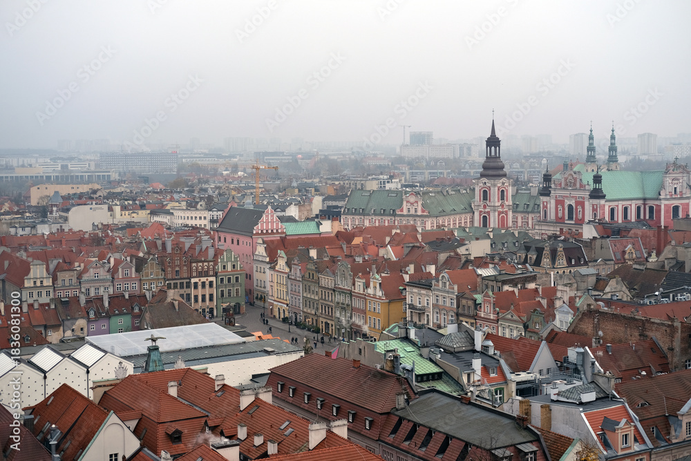 Obraz premium Wide panoramic view of Poznań Old Town with colorful historic buildings, red rooftops and church towers on a misty day.