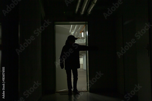 Teenager silhouette at illuminated doorway, dark corridor, hand on doorframe, ambient fluorescent lighting, gritty industrial details, introspective mood, sense of curiosity and threshold moment