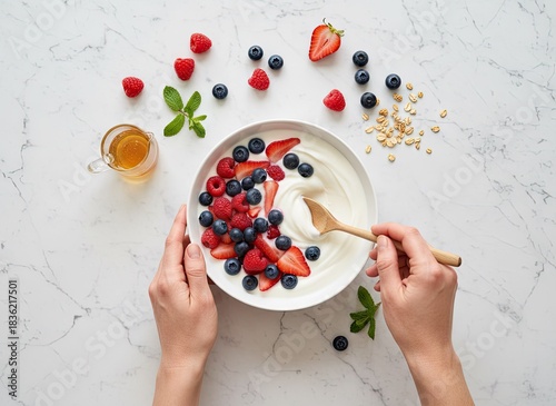 Overhead View Of Hands Preparing A White Yogurt Bowl With Fresh Strawberries Blueberries And Raspberries Sprinkled With Granola And Mint Leaves Beside A Cup Of Tea On A White Marble Surface