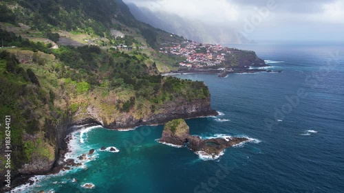Aerial scenic view of Madeira's Seixal resort, perched on rocky cliffs. Colorful houses contrast against lush greenery, with the ocean waves crashing below under a vibrant blue sky