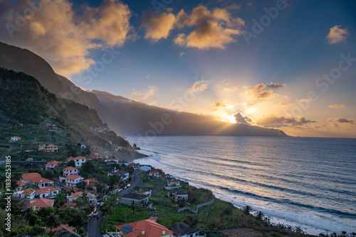 Dramatic aerial view of Madeira's coastline at sunset, showcasing vibrant oranges and blues. The serene ocean, lush mountains, and charming village create a captivating scene