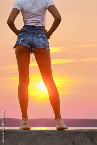 Unrecognizable slender woman in white t-shirt and torn jeans shorts standing on pier at sunset
