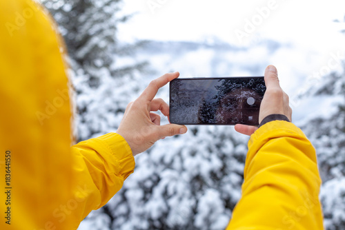 Hands holding a smartphone taking a photo of snowy trees during winter. Close-up view of mobile photography, capturing nature, cold weather and outdoor adventure through technology.