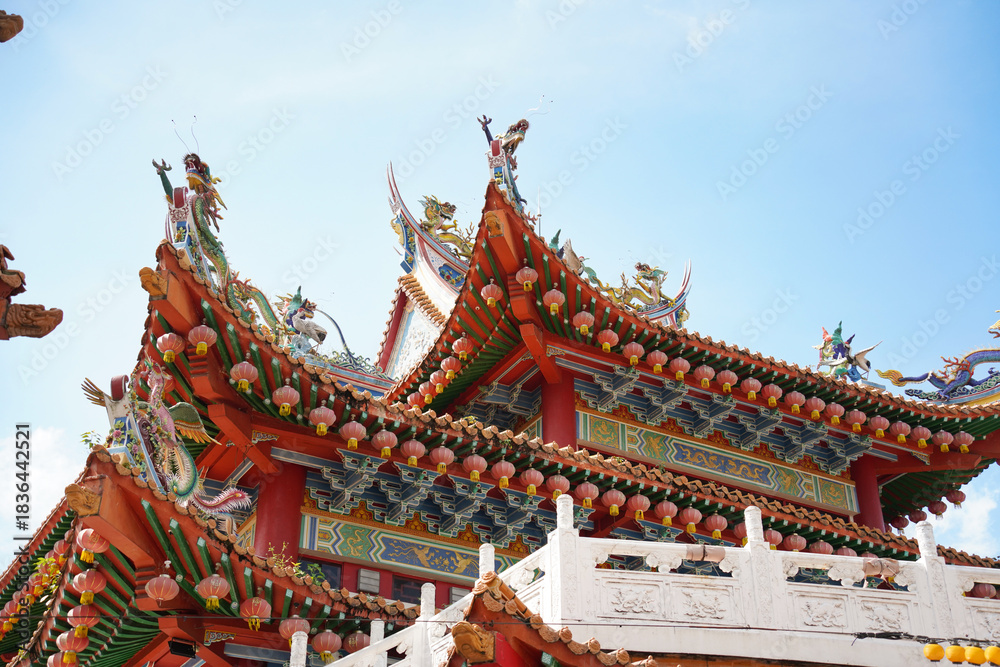 Naklejka premium an ornate, traditional Chinese temple with red, gold, and green colors, adorned with pink lanterns, intricate carvings, and a white balustrade under a clear blue sky.