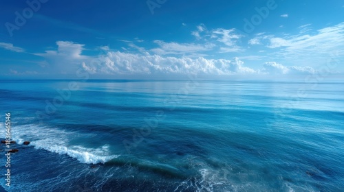 The ocean is calm and blue, with a few clouds in the sky. The water is still, and the waves are small. The sky is clear and bright, and the sun is shining down on the water. The scene is peaceful