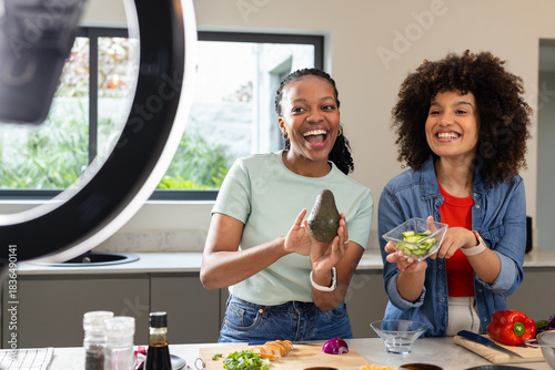 Diverse female friends chopping vegetables under ring light in kitchen, holding avocado, cucumbers