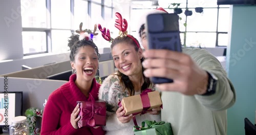 Diverse coworkers reaching for smartphone and capturing holiday selfie with gift boxes at office