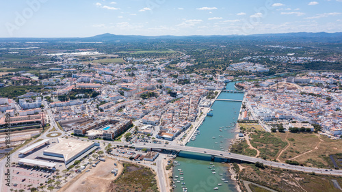 Tavira, a town in Algarve, is seen from the sky with waterways, buildings, and fields. Boats float in the river under bright sunlight and blue sky