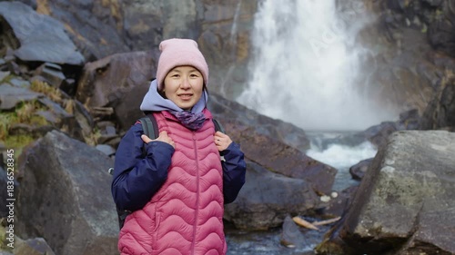 Portrait of a cheerful asian woman hiker with a backpack enjoying the view of a mountain waterfall. This female traveler is standing on a rocky trail, smiling at the camera during her vacation