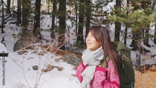 Young smiling asian woman with a backpack enjoying a hike through a beautiful snowy forest, looking around at the serene winter landscape while walking on a trail among pine trees