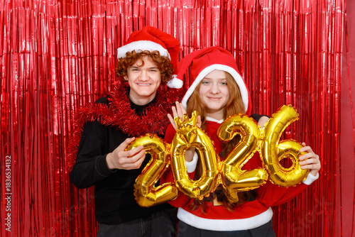 Happy friends in a santa costume holding golden balloons with the shape 2026 on a red background.
