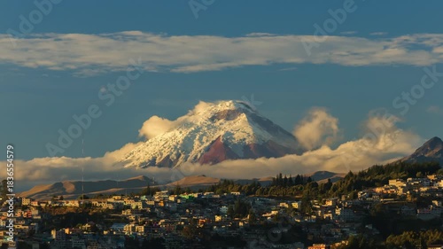Spectacular timelapse of the imposing Cotopaxi volcano, covered in snow and surrounded by clouds, rising above the skyline of Quito, Ecuador.