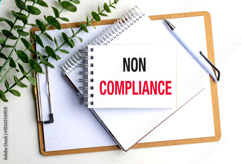 Non compliance concept shown on a notepad with office supplies and plant on white background
