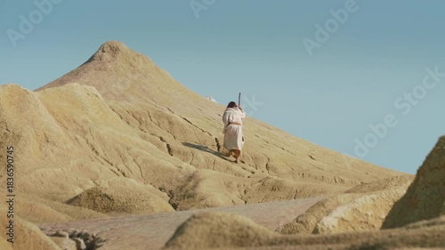 A robed figure walks through a sandy desert holding a wooden staff. Christian and spiritual concept. Slow motion Shot