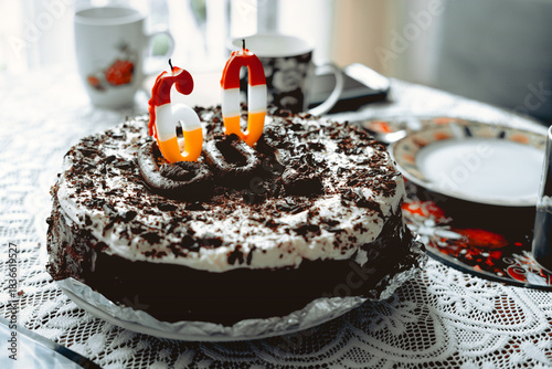 A chocolate cake with whipped cream and a candle with the number sixty on it sits on the table in the living room.