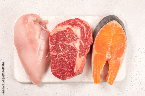 Three pieces of raw meat are placed on a cutting board in a kitchen. The selection includes chicken breast, beef steak, and salmon fillet. The kitchen has a clean and simple look.