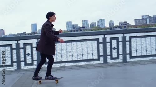 Man in a business suit is rolling on skateboard along embankment, pushing off with his foot, enjoying speed. Guy practicing skateboarding on esplanade. Active, sportive lifestyle, hobby