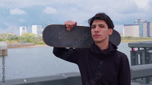 Lonely guy with skateboard on his shoulder is waiting for friends or girl on embankment. Skateboarder enjoying river view, resting, recreating. Boy stands on bridge. Young sportsman walks near river