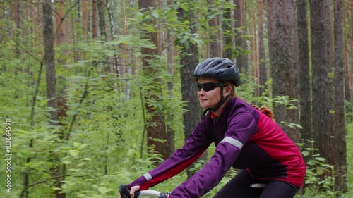 Girl in sports outfit rides cycle through forest. Female cyclist in helmet trains before marathon in woods. Woman practices cycling in nature for health, good mood. Lady moves on bicycle. Side view