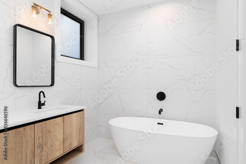 A bathroom detail with a white oak cabinet and white countertop, large format white marble tile walls, and a freestanding tub with a black faucet.
