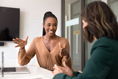 Positive young African manager explaining creative solution for business project to Caucasian partner, talking to female colleague at office table. Two diverse coworkers brainstorming on strategy