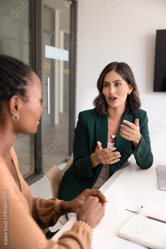 Positive young Latin businesswoman talking to partner at workplace, meeting with consultant for advice, expertise, speaking to colleague, discussing cooperation on project. Vertical shot