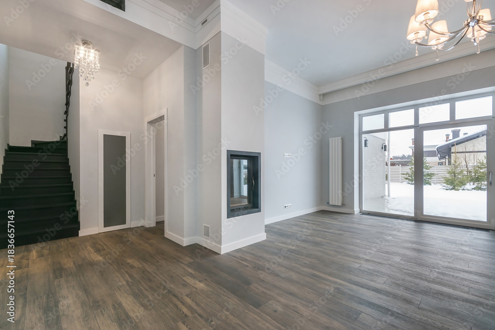 Obraz premium A bright living room with large windows shows a view of snow outside. A staircase leads to the upper level. The fireplace is on the wall as light fixtures hang from the ceiling.