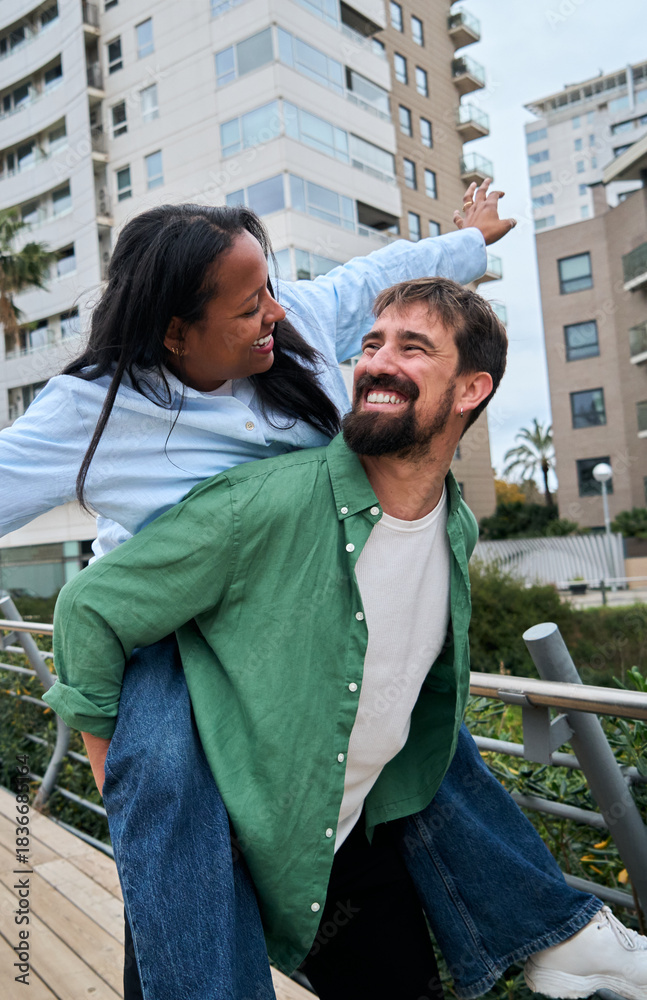 Naklejka premium Vertical. A cheerful couple enjoys a playful piggyback outdoors looking each other, sharing laughter and closeness in a carefree urban moment filled with love, joy, and tenderness.