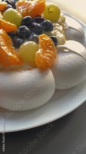 Close-up of a Pavlova meringue dessert topped with creamy filling and fresh berries, including red currants, blueberries and citrus slices. Bright directional light enhances textures and colors.