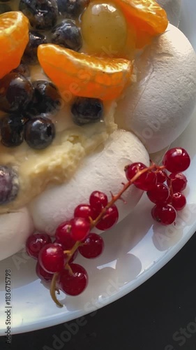 Close-up of a Pavlova meringue dessert topped with creamy filling and fresh berries, including red currants, blueberries and citrus slices. Bright directional light enhances textures and colors.