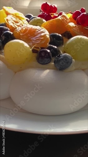Close-up of a Pavlova meringue dessert topped with creamy filling and fresh berries, including red currants, blueberries and citrus slices. Bright directional light enhances textures and colors.