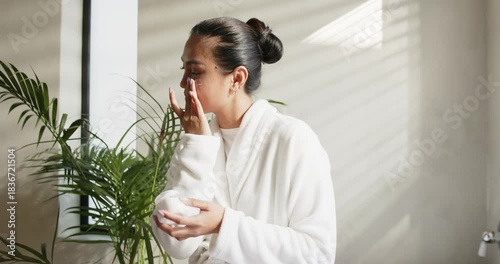 Model opening jar, dipping fingers and massaging face upward by blinds and plant for hydrating skin