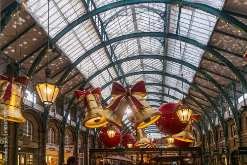 London, UK, 6 December 2025, Giant, illuminated red and gold Christmas bells and globes hang from the arched glass and steel roof of a busy market hall, creating a spectacular festive display.