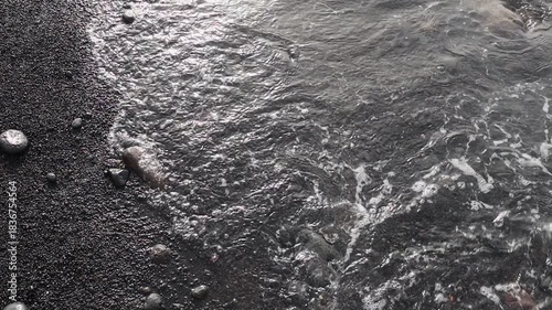 Waves crash and recede into the ocean on a black volcanic pebble beach at sunset.