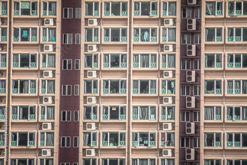 A modern high-rise apartment building with symmetrical windows, green railings, and external air conditioning units, forming a grid-like visual effect.